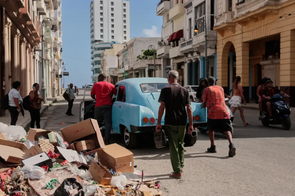 People push a broken-down, classic American car past a pile...