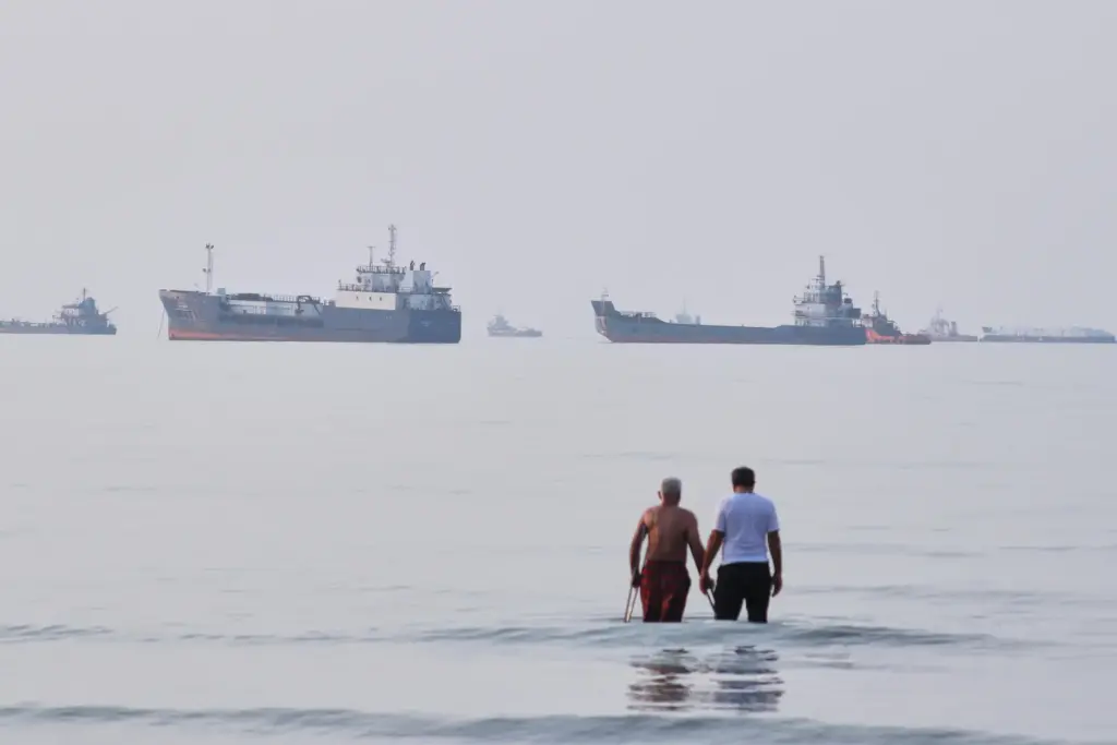 Two men wade in the Strait of Hormuz with vessels anchored in the background.
